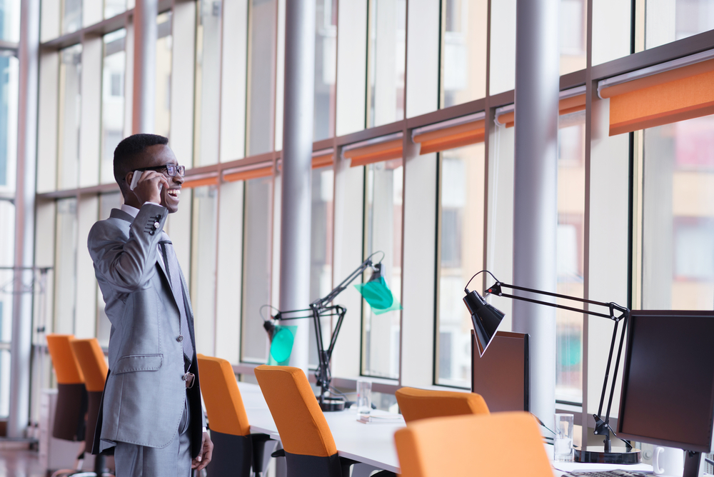 Happy smiling successful African American businessman  in a suit in a modern bright office indoors speel on phone